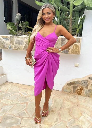 Woman in a vibrant fuchsia wrap dress with spaghetti straps, posing confidently outdoors with gold accessories and sandals.