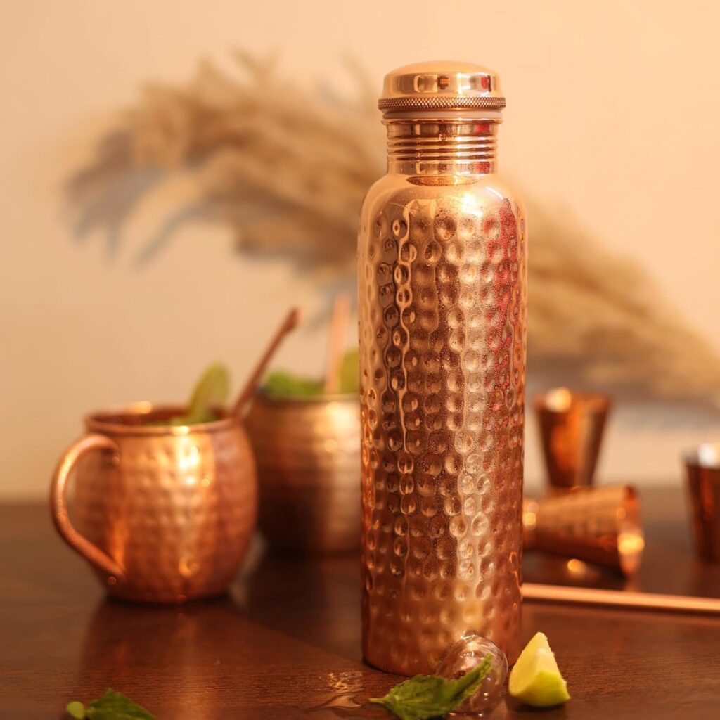 Hammered copper water bottle with matching mugs and cocktail tools on wooden table with lime, mint, and ice.