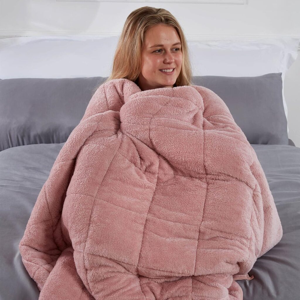 Woman wrapped in a soft pink Brentfords teddy weighted blanket, sitting on a bed with a peaceful expression