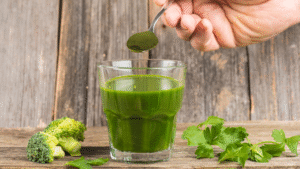 Hand scooping spirulina or wheatgrass powder into a glass of green juice with broccoli and parsley