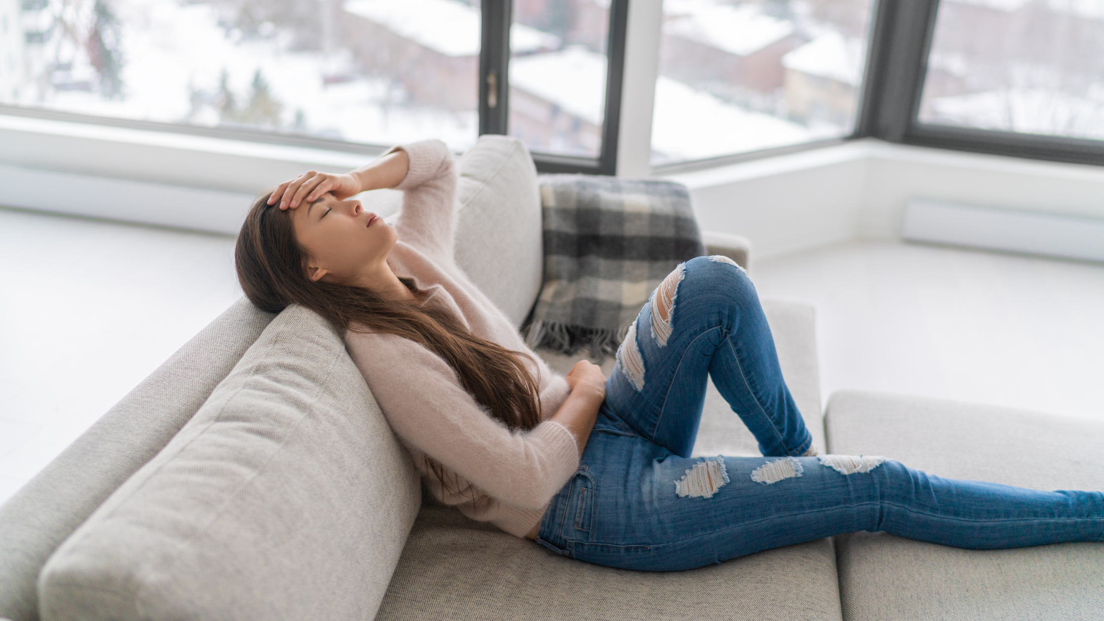 Woman in distress holding her forehead while lying on a modern grey sofa, expressing emotional exhaustion and trauma recovery