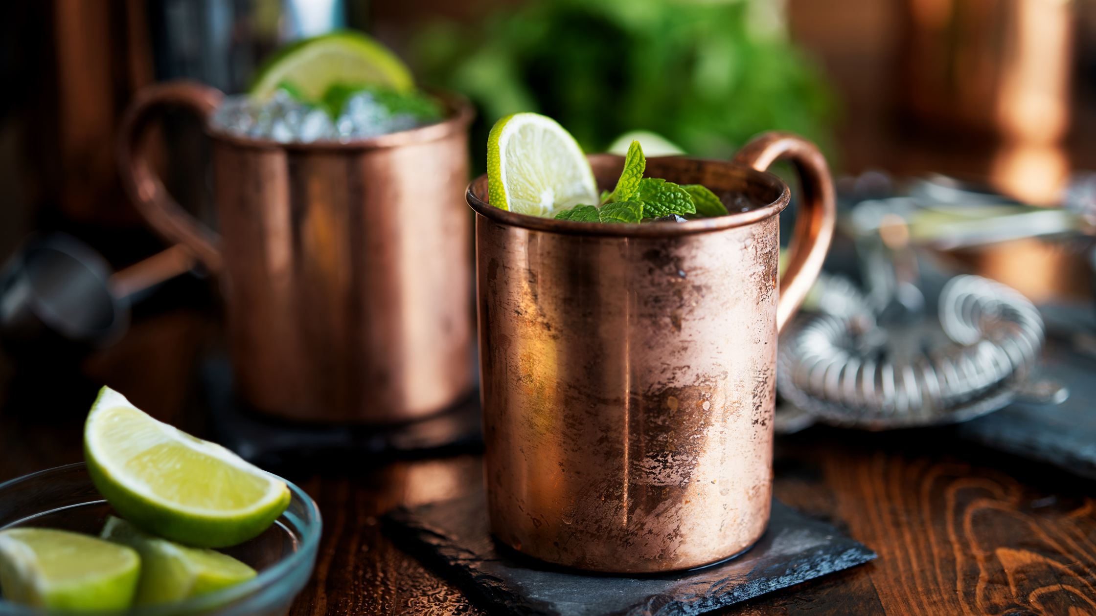 Alt Text: Copper mugs filled with a refreshing lime and mint drink on a dark wooden table, surrounded by lime wedges and bar tools.