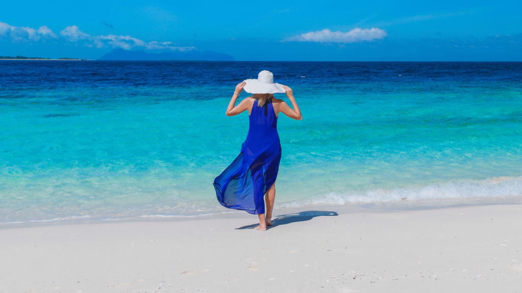 Woman in a flowing cobalt blue dress walking along a turquoise beach, facing the ocean with a white sunhat, embodying peace, clarity, and self-expression