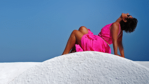Woman in a vibrant fuchsia wrap dress reclining on white sand under a blue sky, embodying elegance, softness, and emotional strength