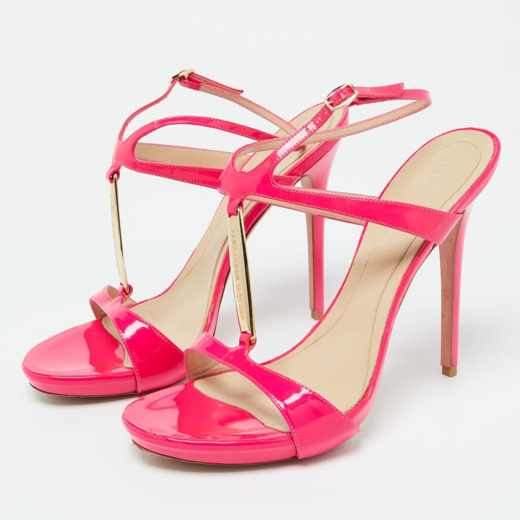 Close-up of Alexander McQueen fuchsia pink patent leather high-heeled sandals with gold bar detail, placed against a white background