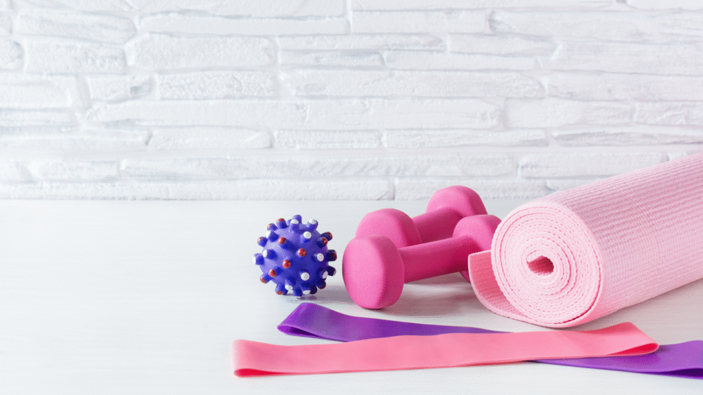 Flat lay of pink and purple resistance bands, yoga mat, dumbbells, and massage ball on white wooden surface against textured white wall