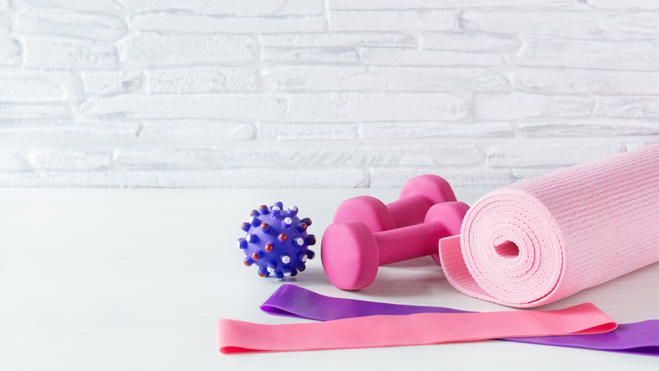 Flat lay of pink and purple resistance bands, yoga mat, dumbbells, and massage ball on white wooden surface against textured white wall
