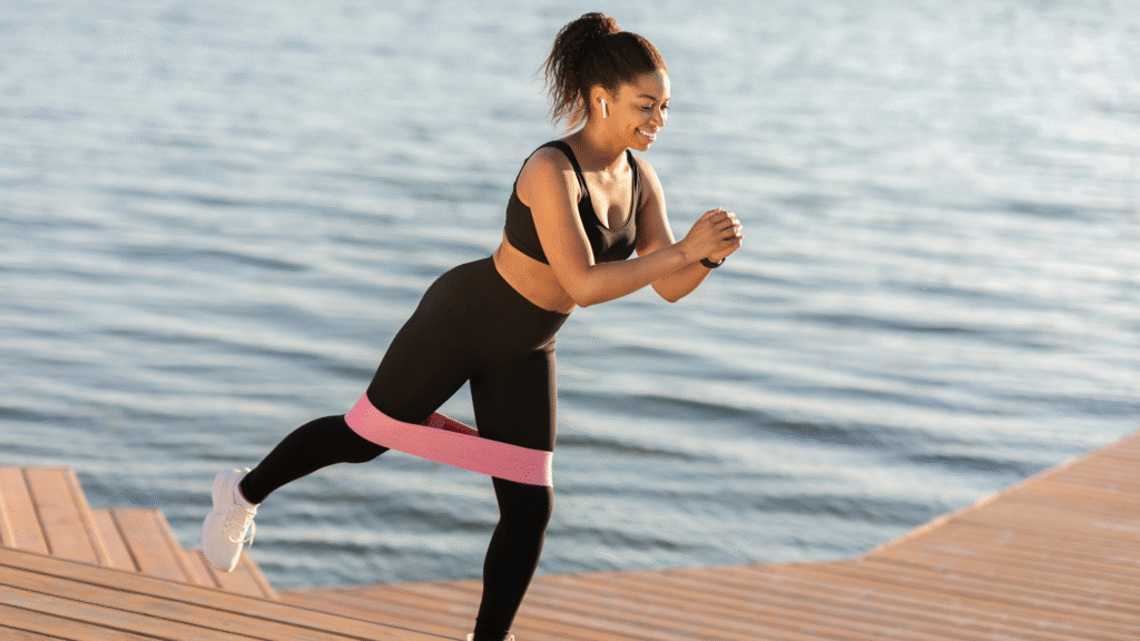 Woman exercising with pink resistance band outdoors on wooden dock by the water, balancing in athletic pose